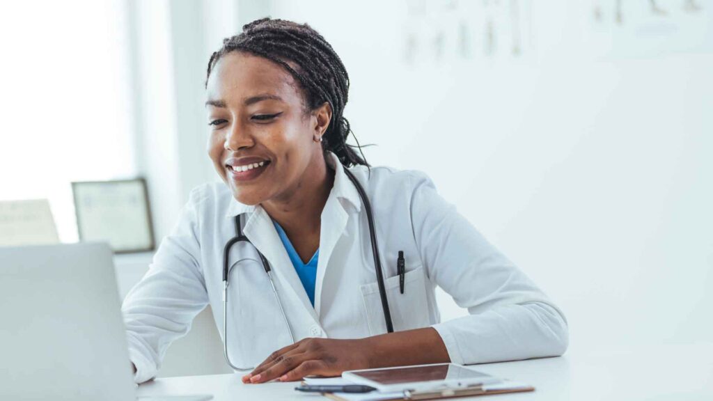 Concentrated afro american woman doctor using laptop while sitting at workplace in medical clinic